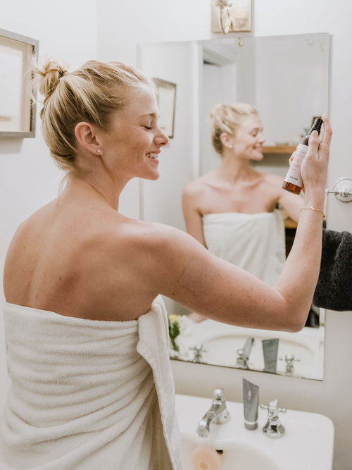 Smiling woman in a towel applying skincare in a bright bathroom mirror