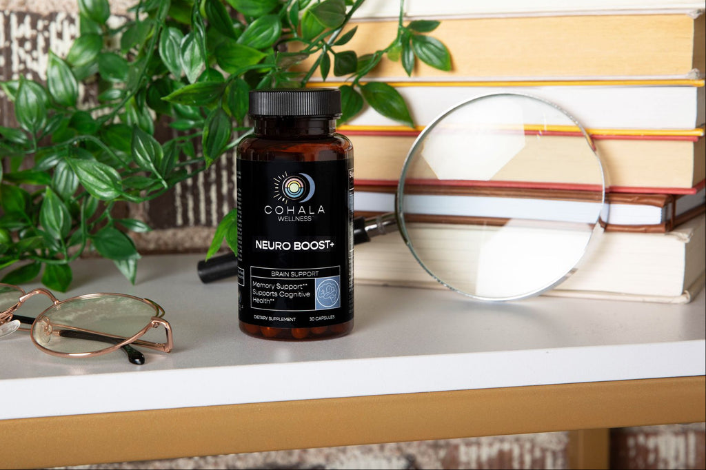 COHALA Wellness Neuro Boost+ supplement bottle on a shelf with books, glasses, a magnifying glass and greenery